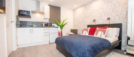 A modern bedroom with a large bed, a small kitchenette, and a bathroom visible through an open door.
