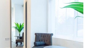 A room with a grey armchair, a round table, a large mirror, and a window with a view of greenery.
