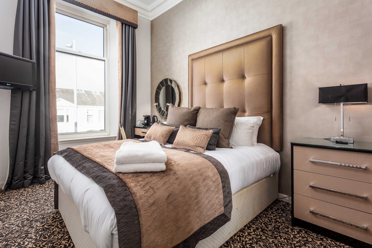 A hotel room with a large bed, a bedside table with a lamp, a TV, and a window with curtains.