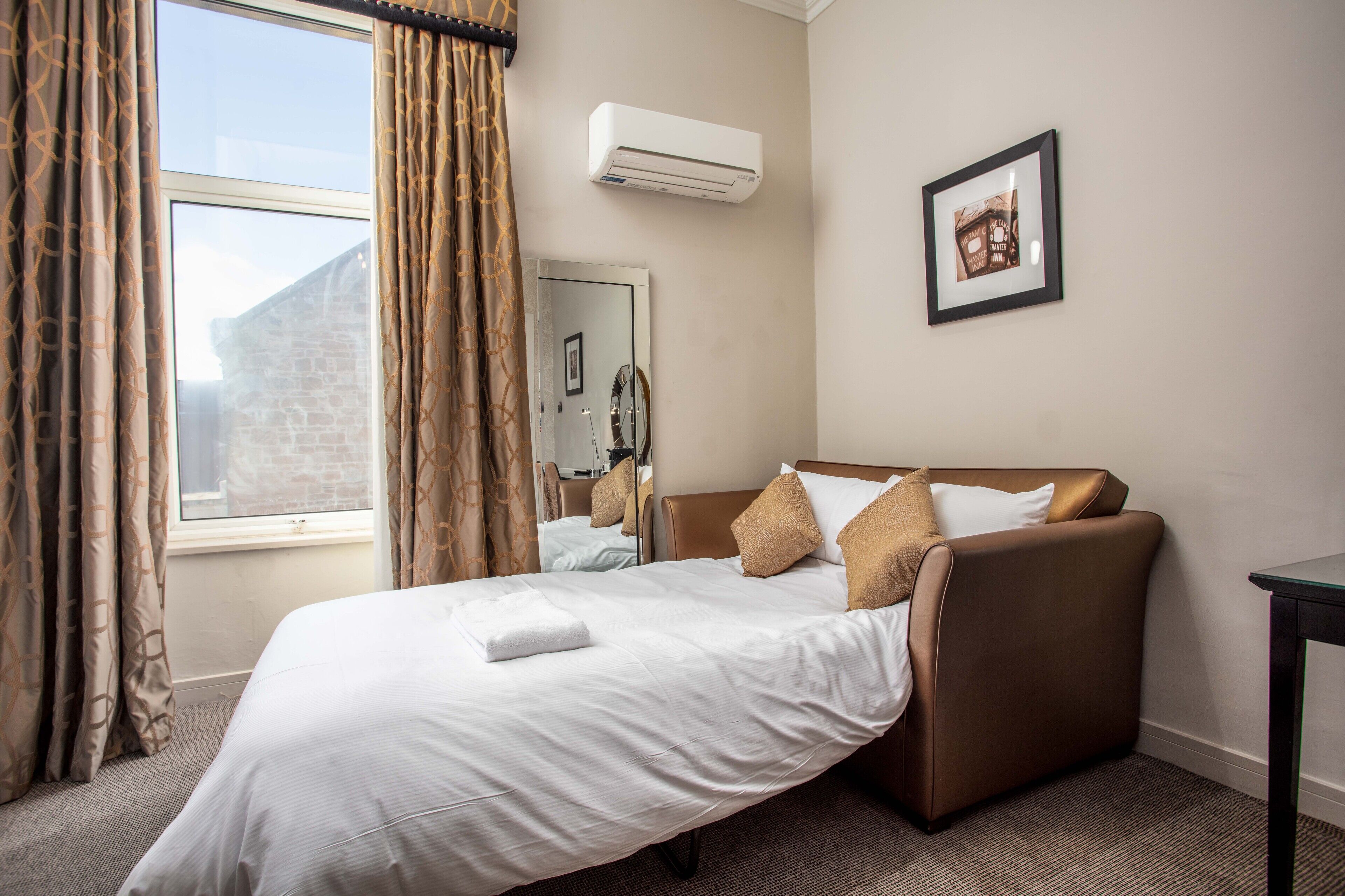 A hotel room with a bed, a window with curtains, an air conditioning unit, a framed picture on the wall, and a carpeted floor.