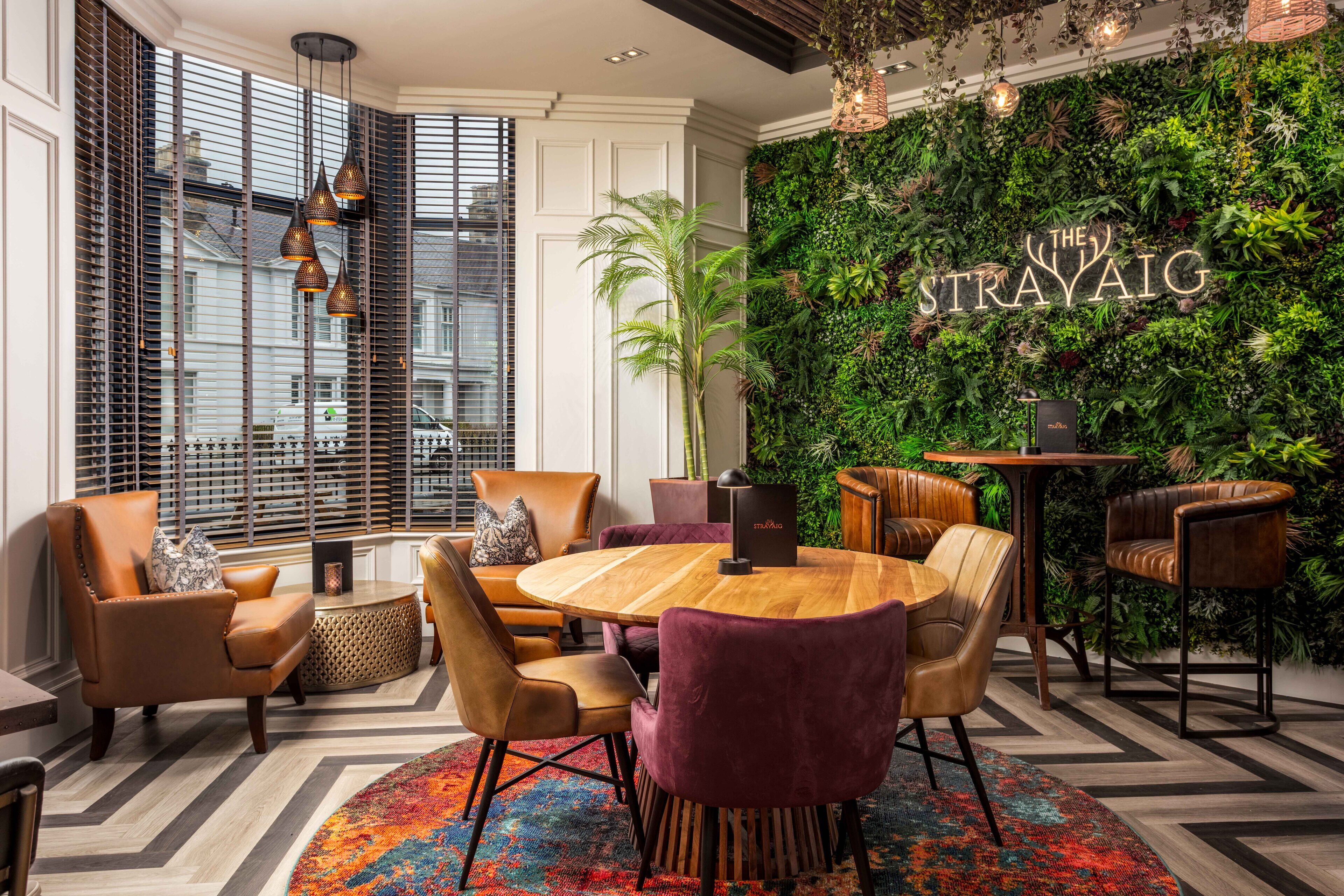 Een moderne lounge met een ronde houten tafel, lederen fauteuils en een groene wand met het bord 'The Strayaig'.