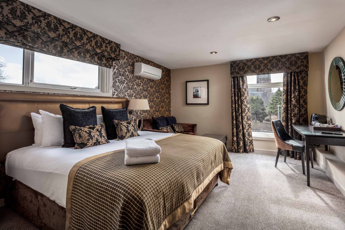 A hotel room with a large bed, a desk, and a chair. There is a window with curtains, a wall-mounted air conditioner, and a framed picture on the wall.
