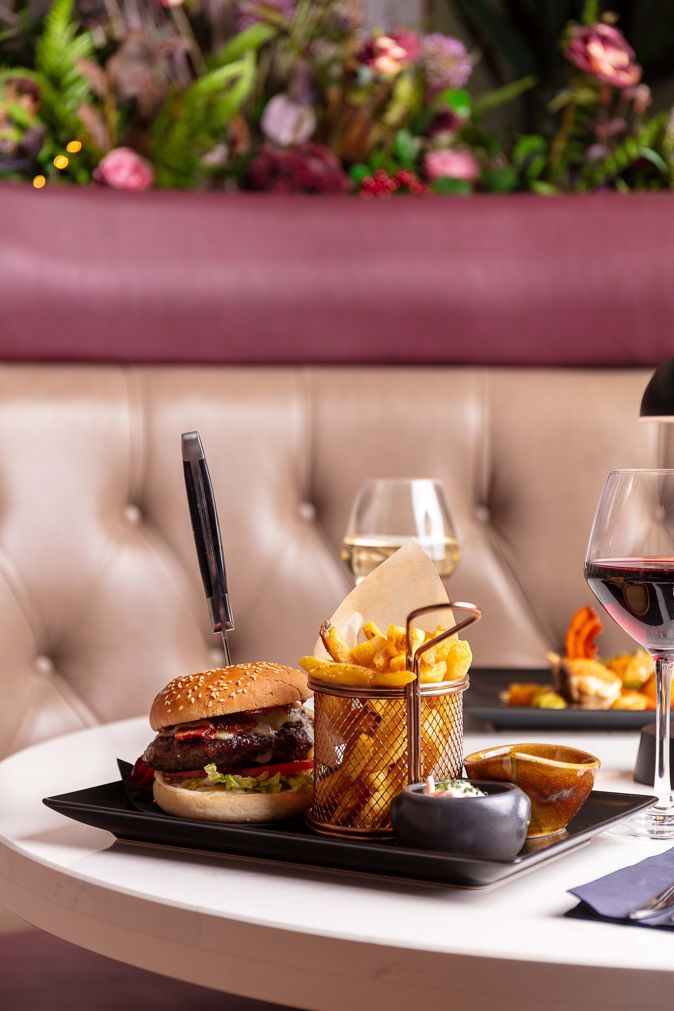 Una mesa de comedor con una hamburguesa, una cesta de papas fritas, un tazón pequeño y una copa de vino tinto.