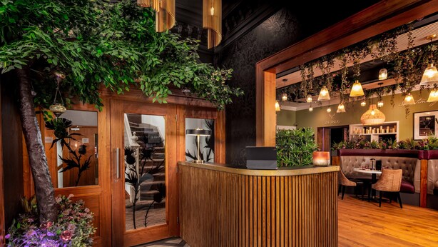 Zona de recepción con mostrador de madera, escalera y área de descanso con banco acolchado y mesa.