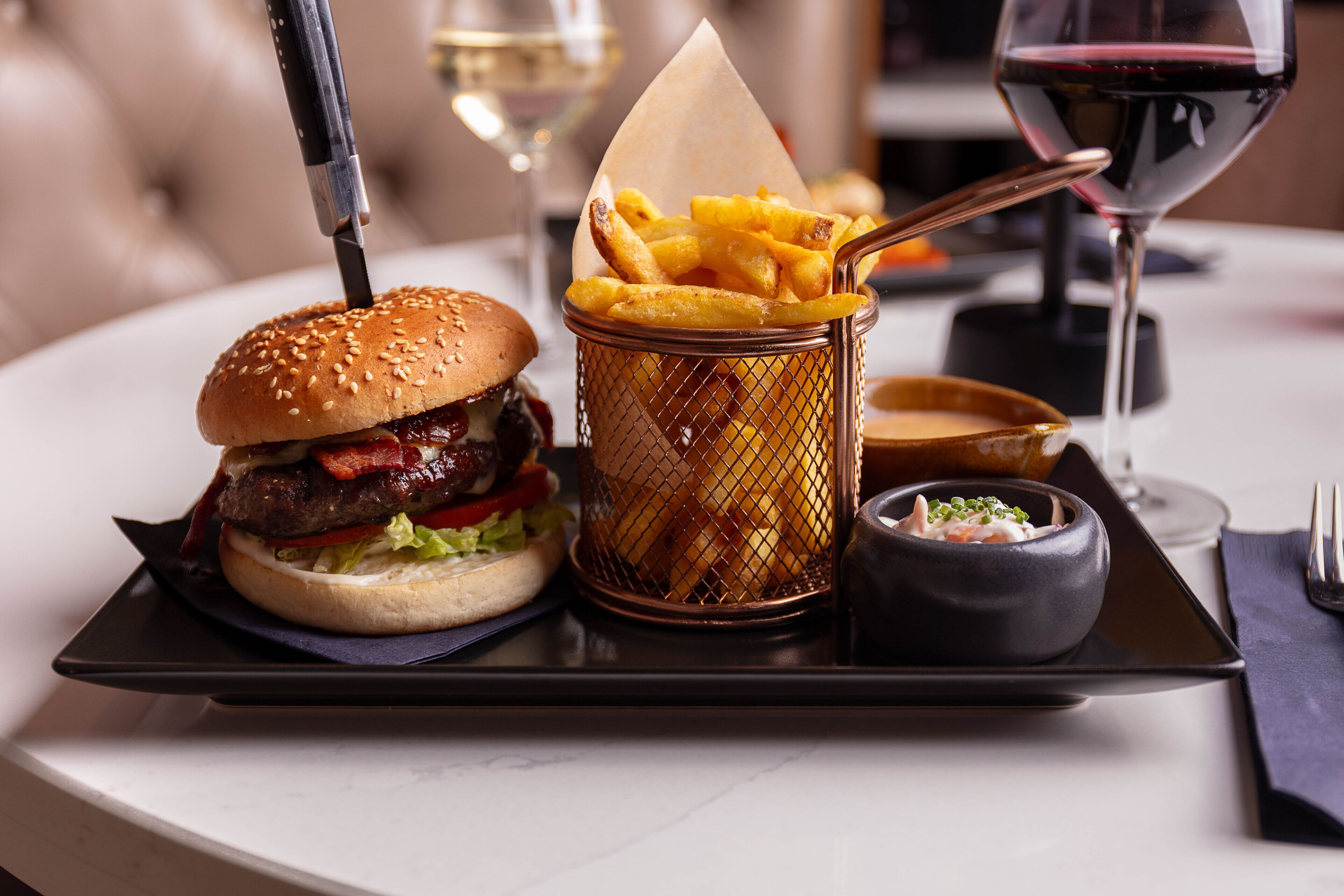 A meal tray with a burger, fries, and a small bowl of sauce, accompanied by a glass of red wine.