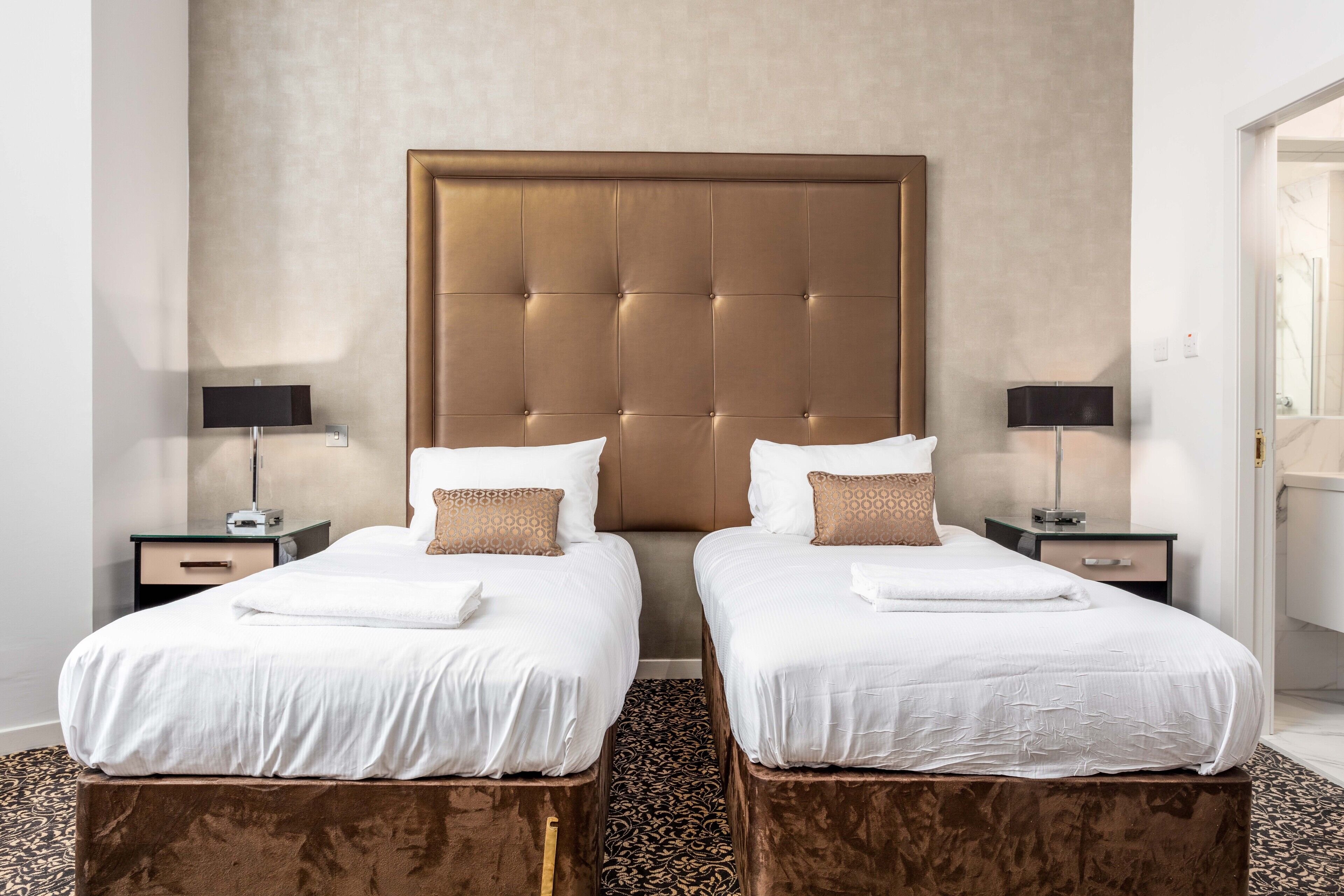 A hotel room with two beds, a large tufted headboard, bedside tables with lamps, and a patterned carpet.