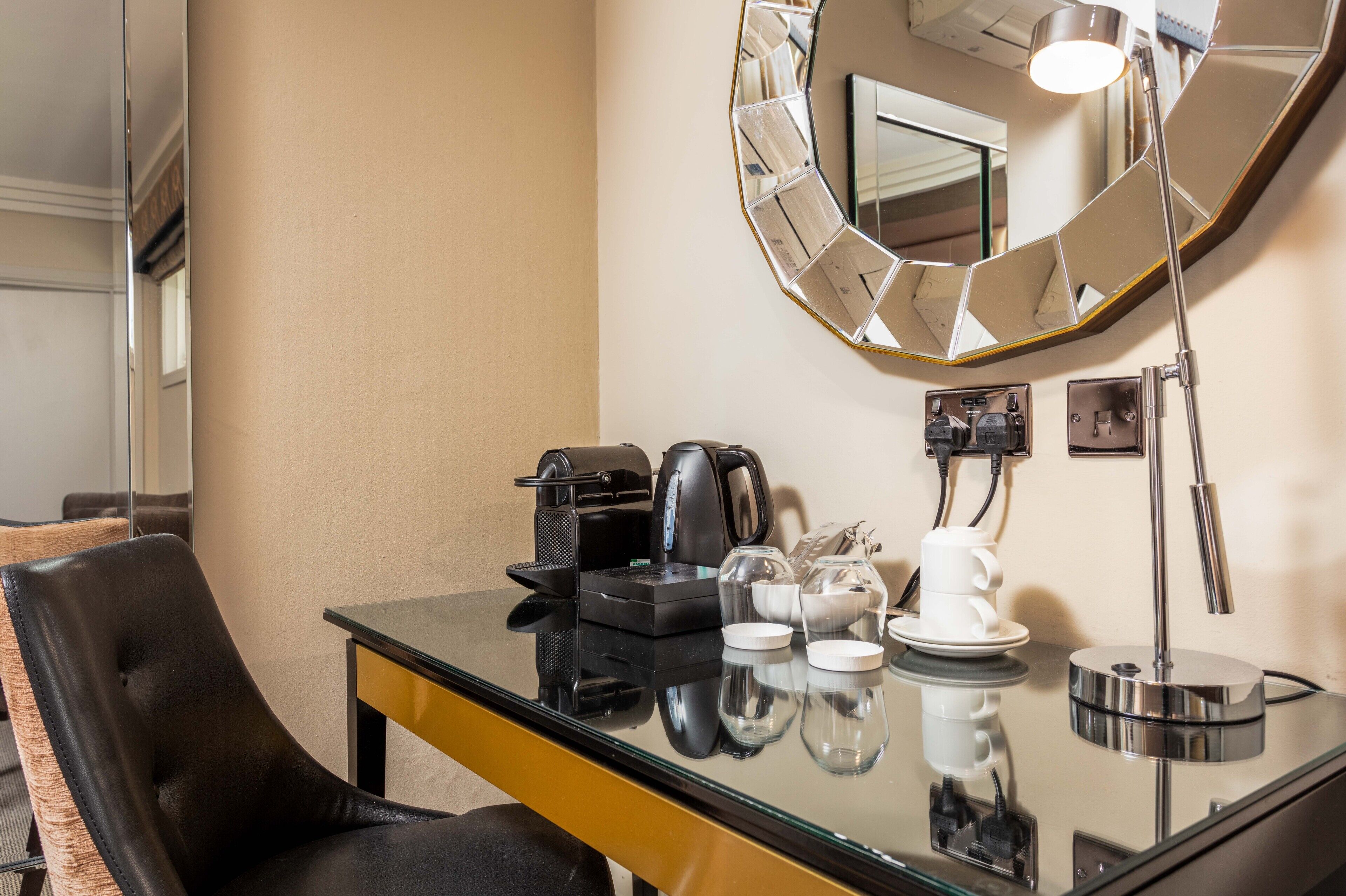 A hotel room with a glass table, a black leather chair, a coffee maker, a kettle, a mirror, and a lamp.
