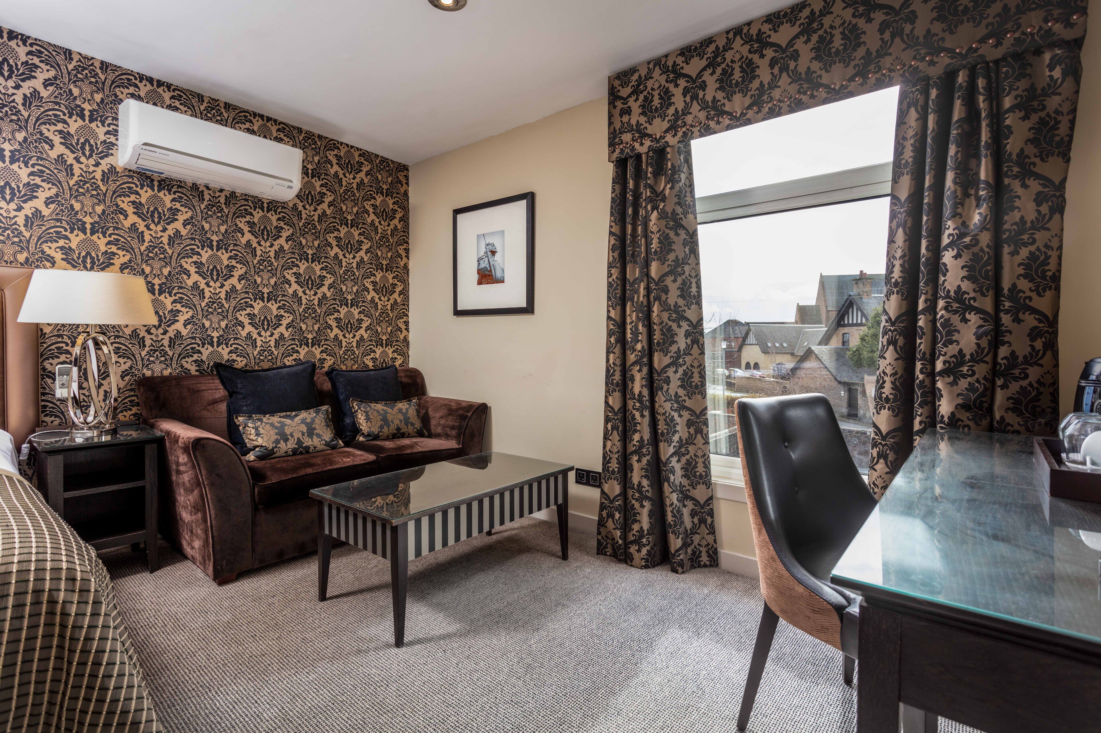A hotel room with a patterned wallpaper, a brown sofa with cushions, a glass coffee table, a dining table with a chair, and a large window with curtains.