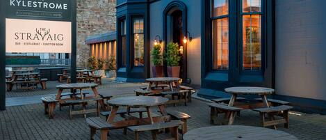 An outdoor seating area with wooden tables and benches, a sign for The Kyleystone, and a building with blue and white walls.
