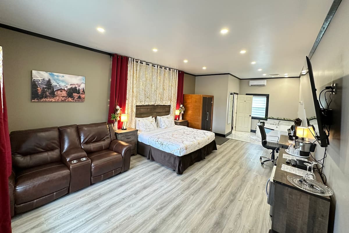 Una habitación de hotel moderna con cama, sofá, escritorio con computadora, y baño visible a través de una puerta abierta.