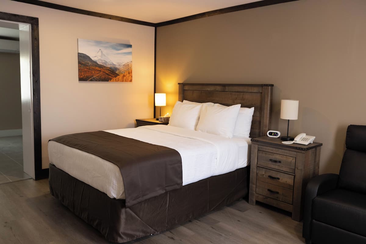 Habitación de hotel con una cama grande, un cabecero de madera, una mesita de noche con lámpara, teléfono y una silla.