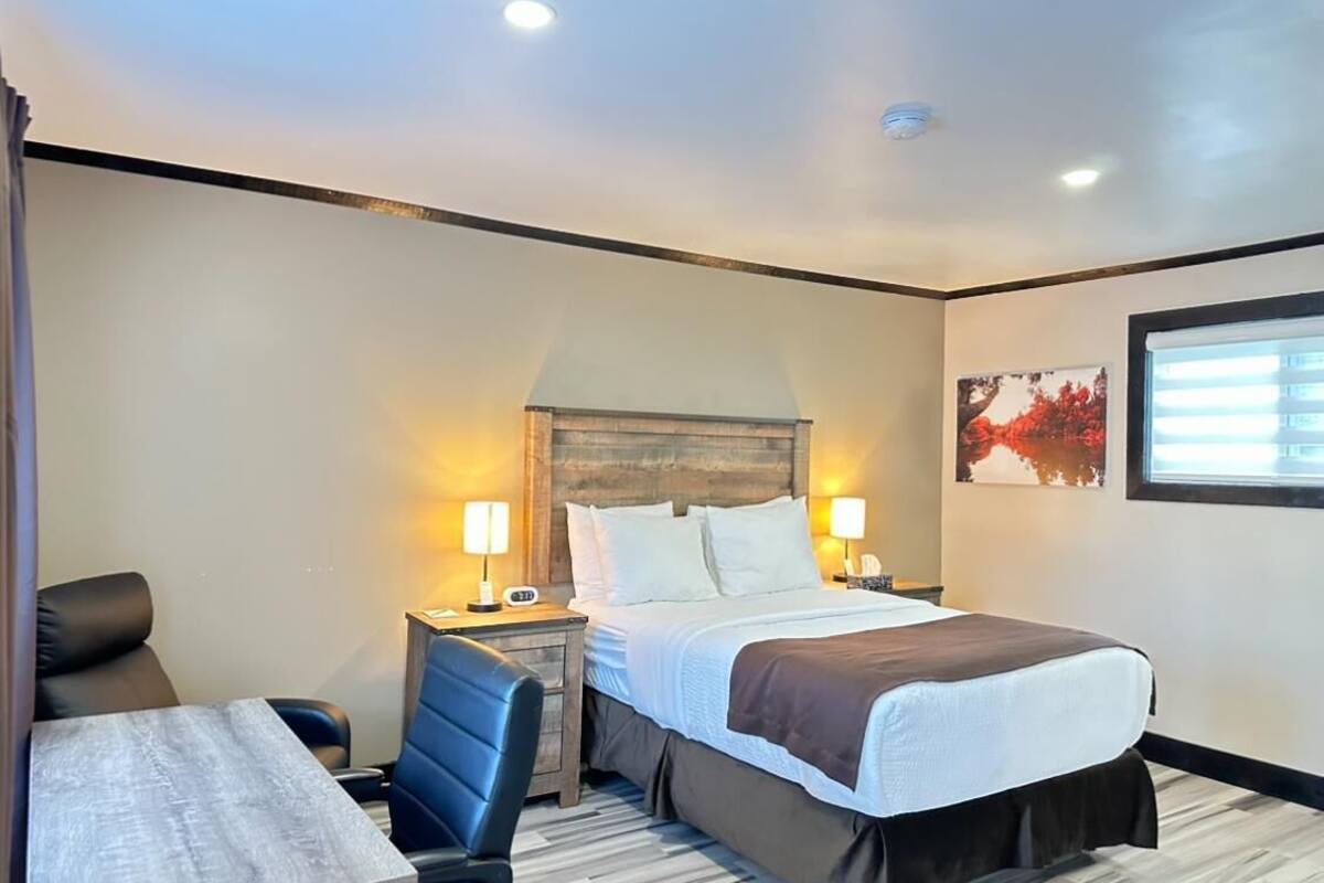 Habitación de hotel con cama, escritorio, dos sillas, mesita de noche con lámpara y ventana con persianas.