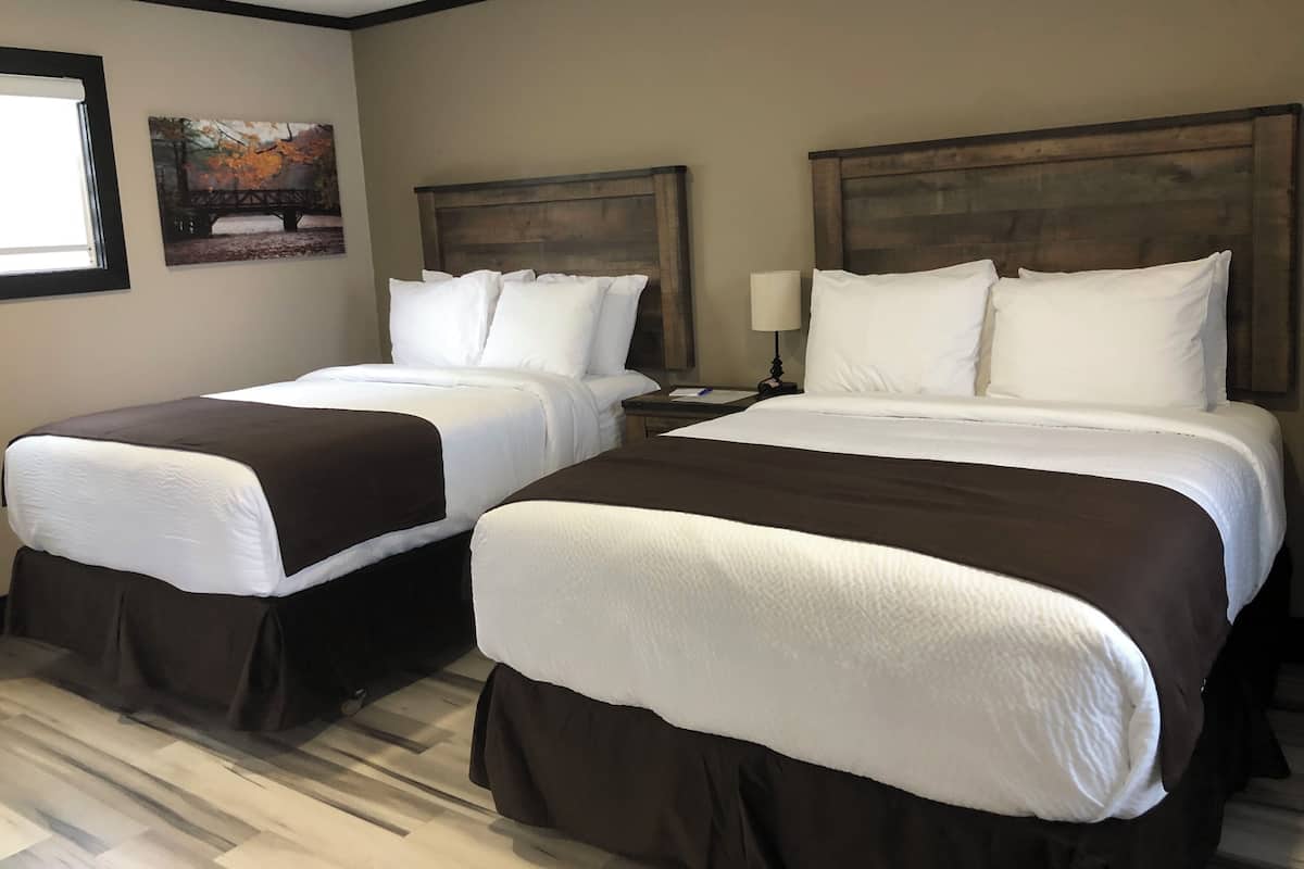 Habitación de hotel con dos camas, cada una con ropa blanca y cabeceros marrones. Se ve una ventana a la izquierda y un cuadro en la pared sobre la ventana.