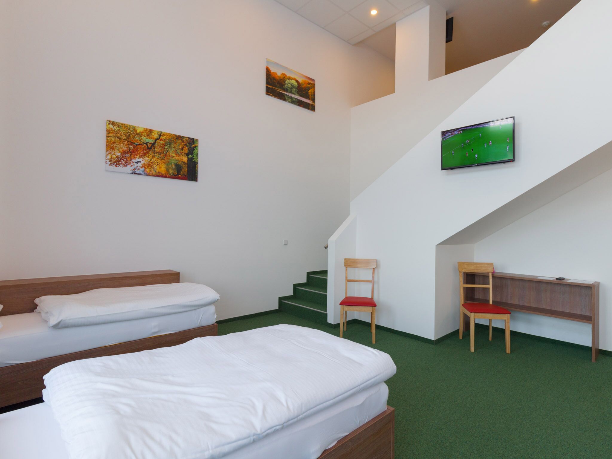 A hotel room with two beds, a TV mounted on the wall, a staircase, and a chair.