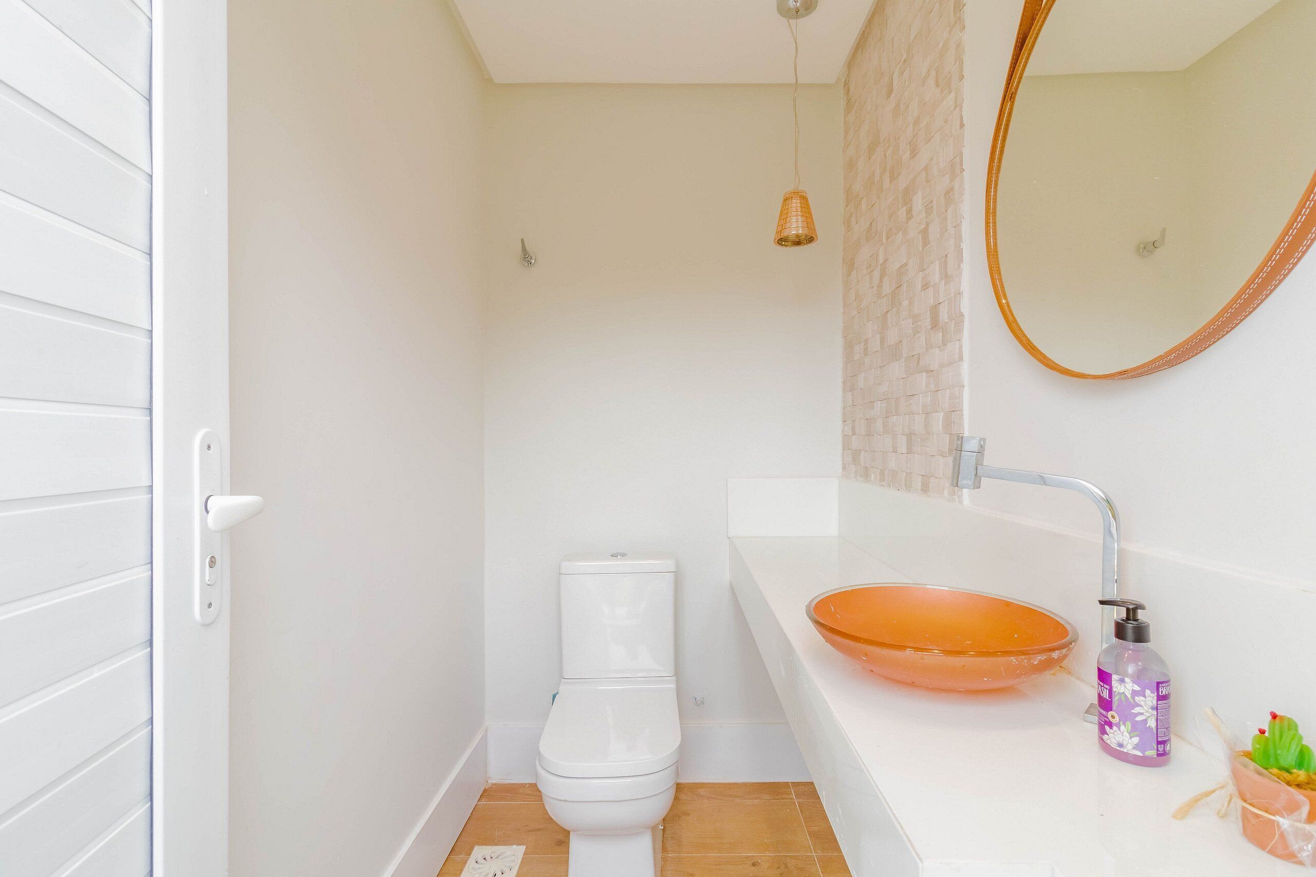 Un baño con un inodoro blanco, un lavabo blanco con un lavamanos naranja y un dispensador de jabón morado, además de una lámpara colgante.