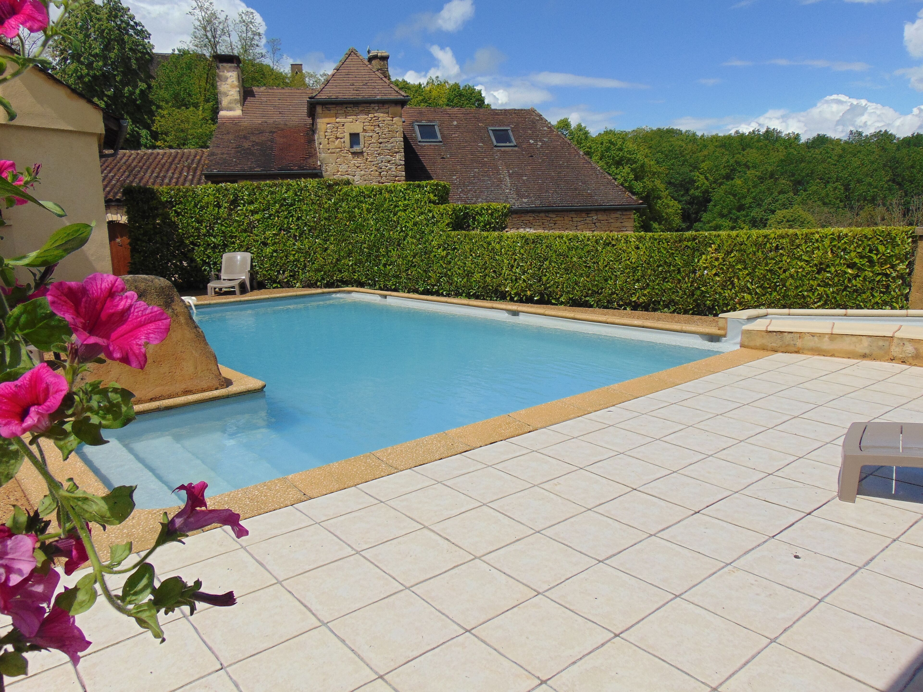 A swimming pool with a tiled deck, surrounded by a hedge and a stone building in the background.