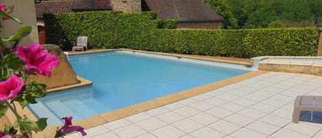 A swimming pool with a tiled deck, surrounded by a hedge and a stone building in the background.
