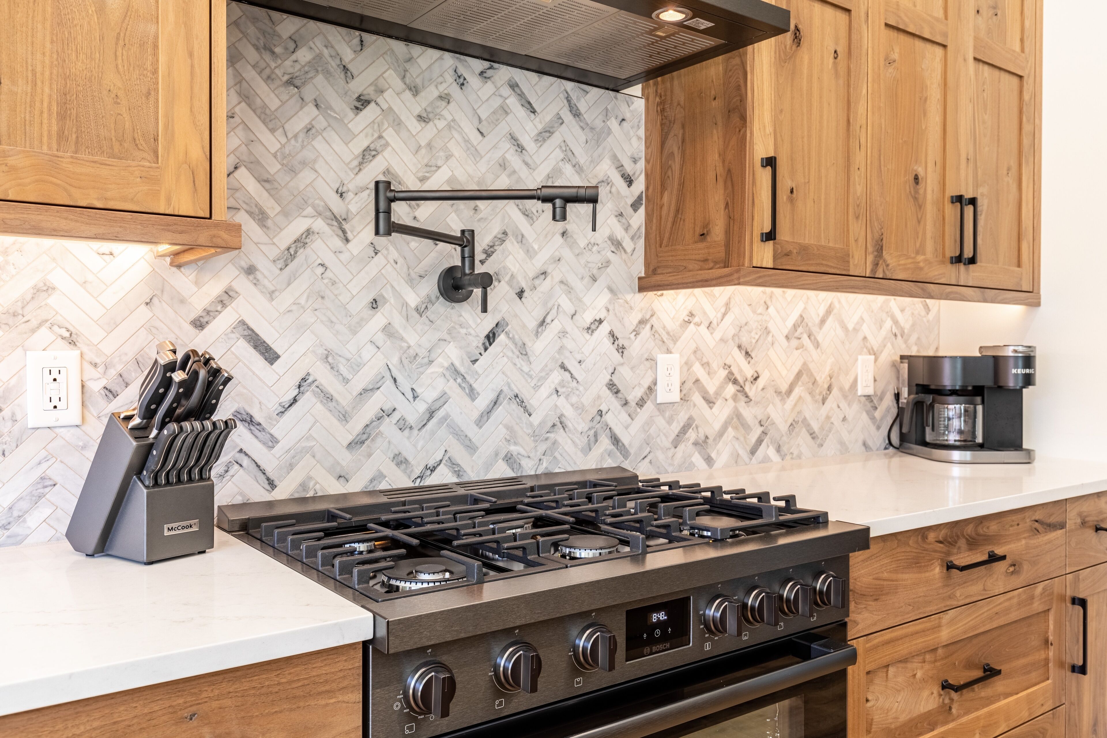 Een keuken met een backsplash in een herringbone-patroon, houten kastjes, een roestvrijstalen afzuigkap en een ingebouwd gasfornuis.