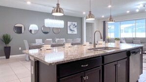 A modern kitchen with a granite countertop, dark cabinetry, and pendant lighting. There are three round mirrors on the wall, a potted plant, and a dining area with a table and chairs. - Luxurious Pool Home With Private Pool-2061lc 8 Bedroom Home by RedAwning (Davenport)