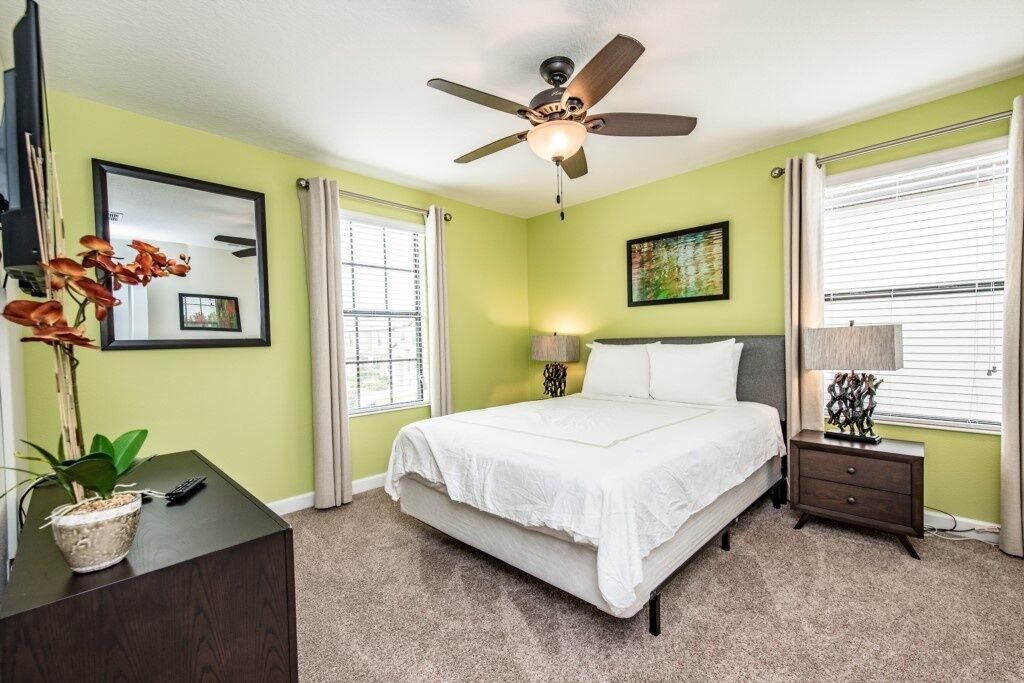 A bedroom with a bed, a ceiling fan, a bedside table with a lamp, a dresser, and a window with curtains.