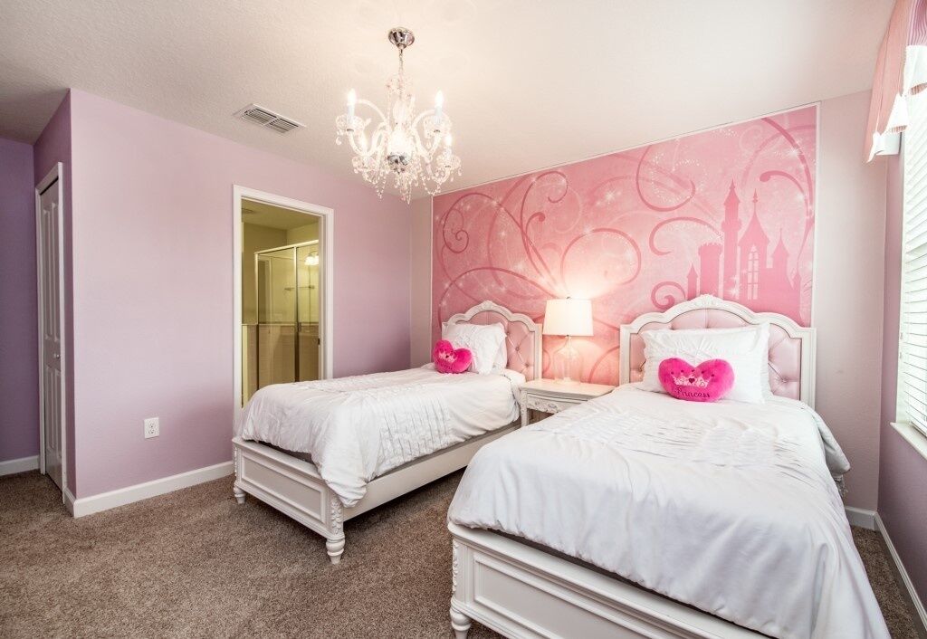 A bedroom with two white beds, a chandelier, and a pink wall with a castle design.