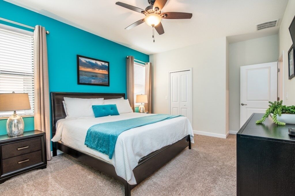 A bedroom with a bed, nightstands, a ceiling fan, a painting, and a desk with a plant.