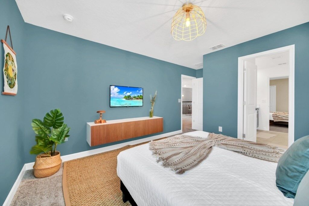A bedroom with a bed, a television, a plant, a console table, and a door leading to another room.