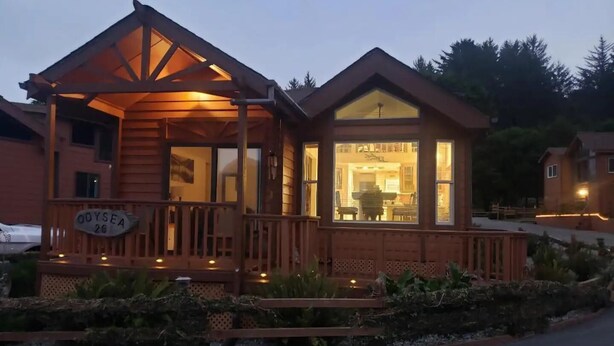 A wooden cabin with a porch, a visible sign reading "ODYSEA 26", and a window showing a dining area inside. - Odysea Modern Ocean Cabin 26 w/ Jacuzzi & Modern Decor (Private Beach Owned by White Rock Resort) (Smith River)