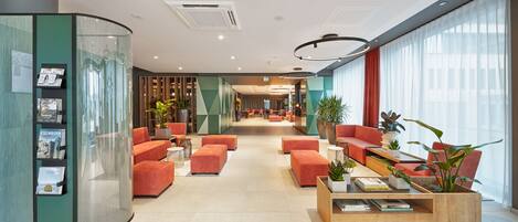 A modern lobby with a curved reception desk, orange seating, and a coffee table with books and plants.