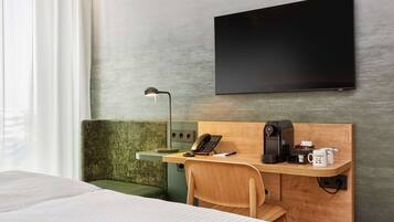 A hotel room with a bed, a desk with a telephone, a coffee maker, and a flat-screen TV mounted on the wall.