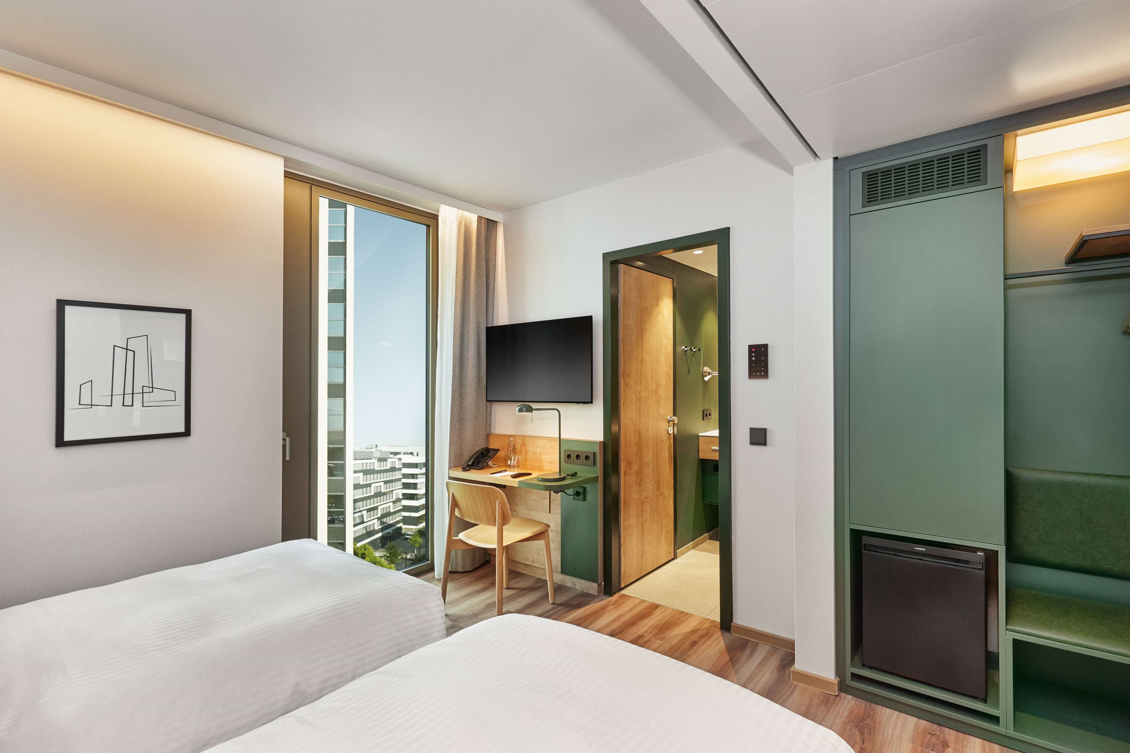 Habitación de hotel con dos camas, un escritorio con silla, televisor, armario y vista a la ciudad.