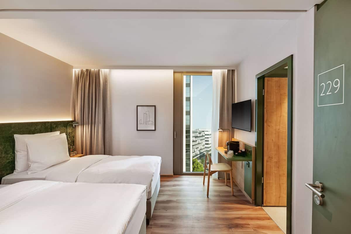 Ein Hotelzimmer mit zwei Betten, einem Schreibtisch, einem Fernseher und einem Balkon mit Blick auf die Stadt.