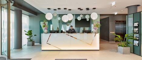 A modern hotel reception area with a glass desk, a receptionist, and a digital screen.