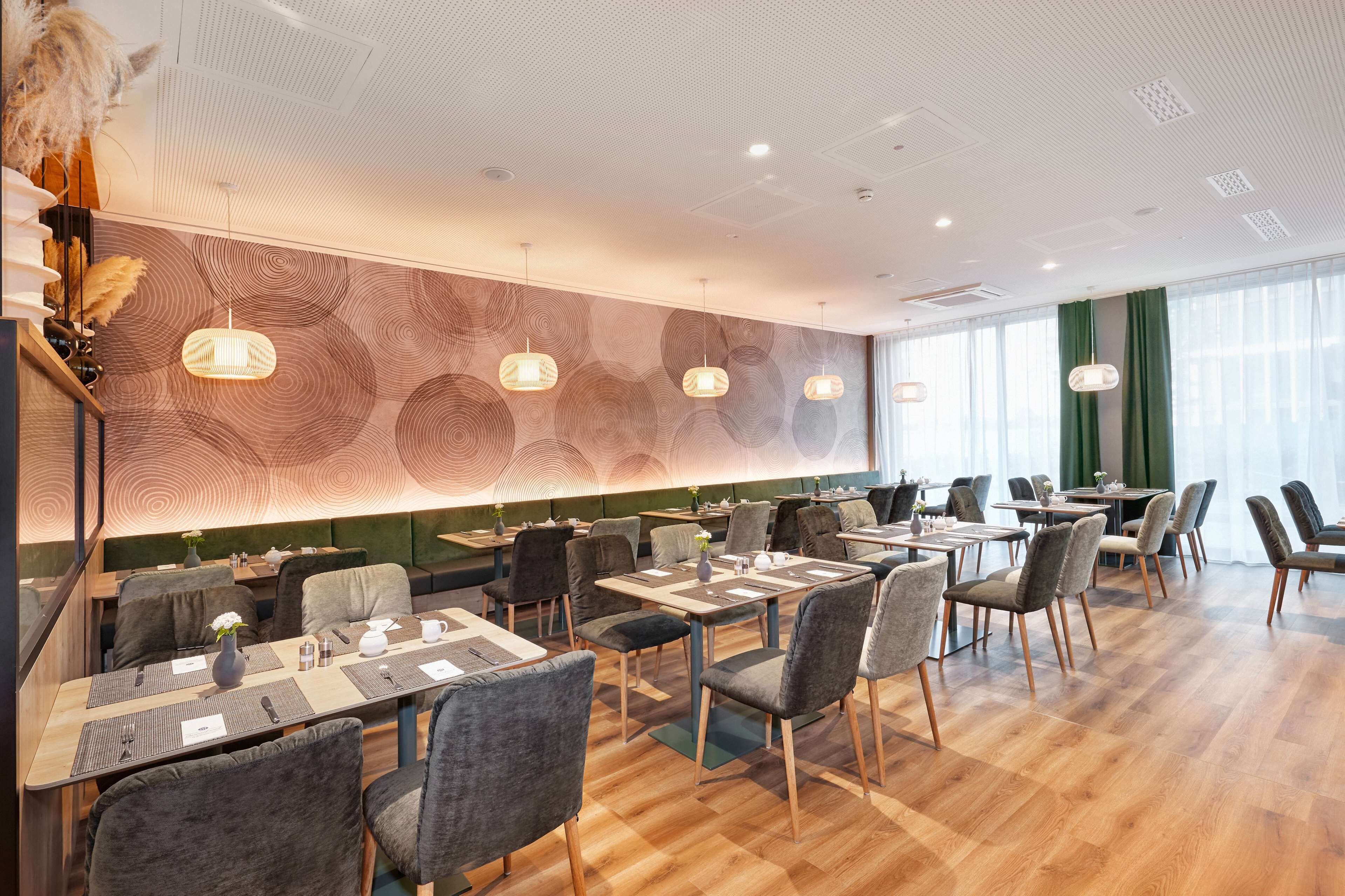 Un restaurant moderne avec un plancher en bois, des chaises à revêtement gris et des luminaires suspendus. Les tables sont dressées avec des sets de table, des couverts et des verres à eau.