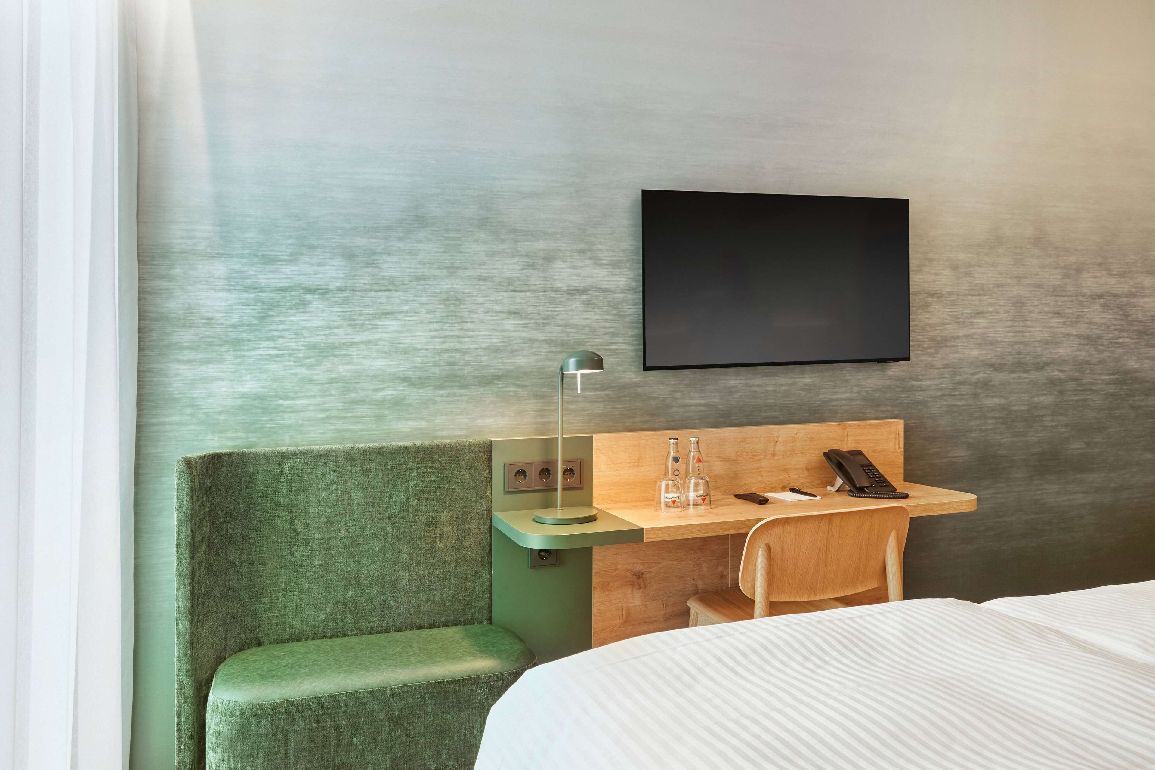 Camera d'albergo moderna con una testiera imbottita verde, una scrivania in legno con una lampada, una TV a schermo piatto e un letto con copripiumino a righe.