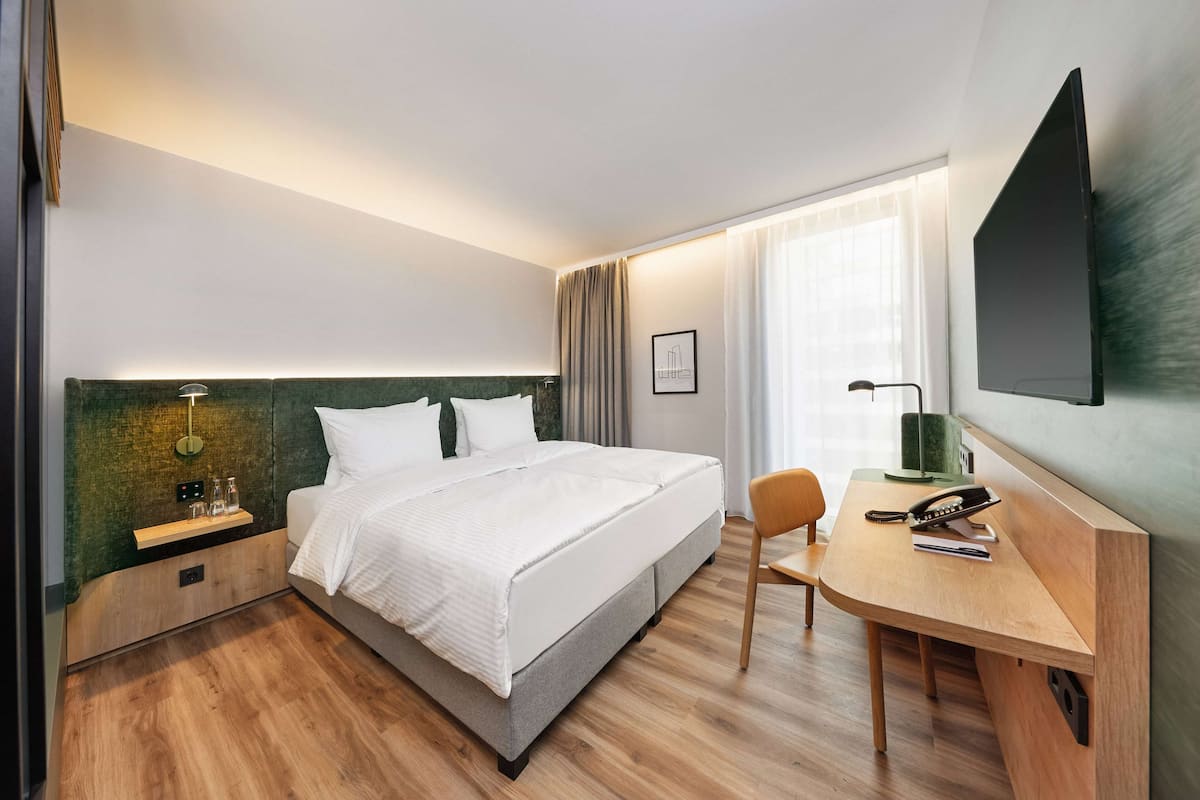 Ein modernes Hotelzimmer mit einem großen Bett, einem Schreibtisch mit Stuhl, einem Flachbildfernseher und einem Fenster mit Vorhängen.