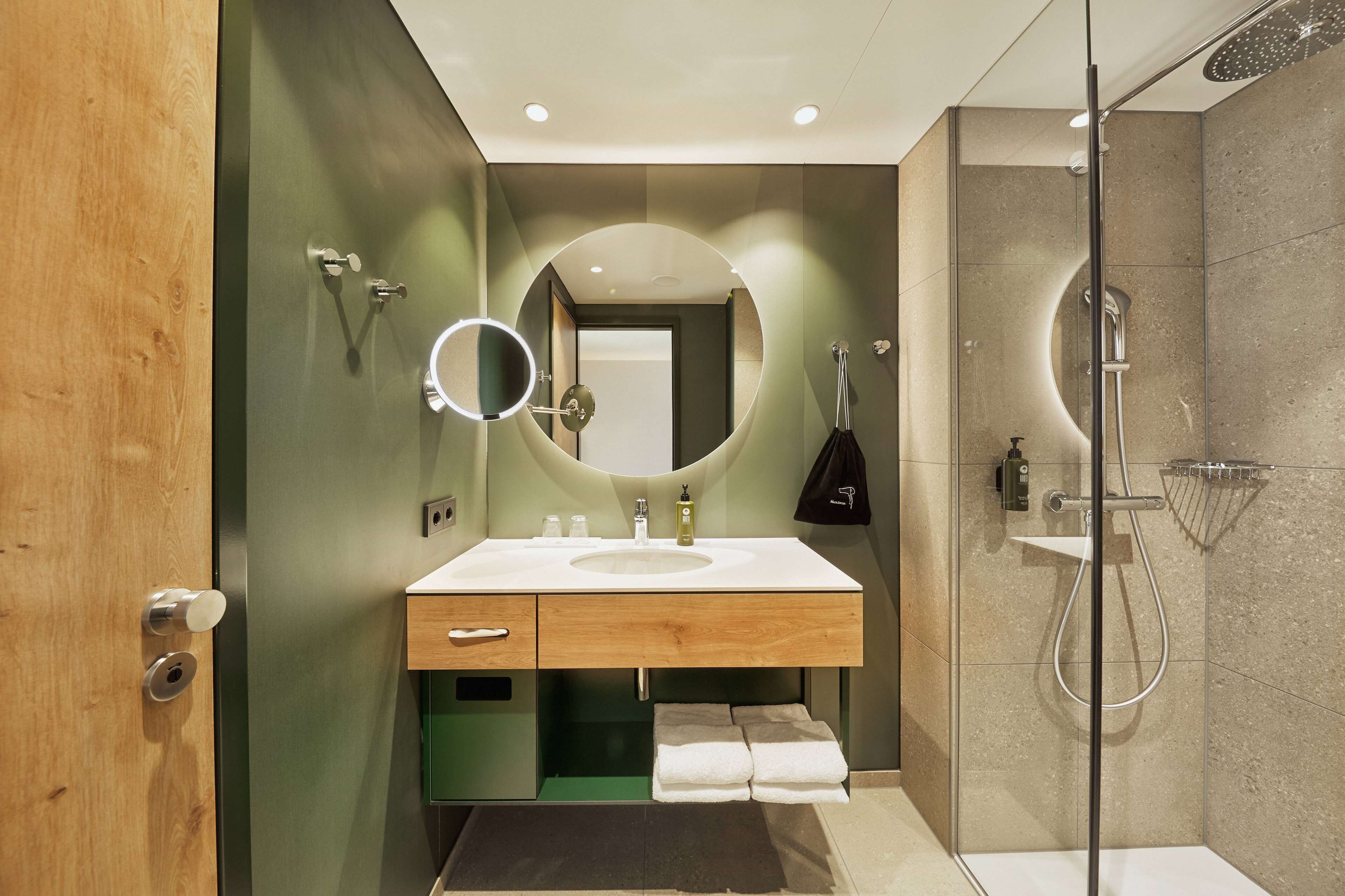A modern bathroom with a wooden vanity, a round mirror, and a glass-enclosed shower.