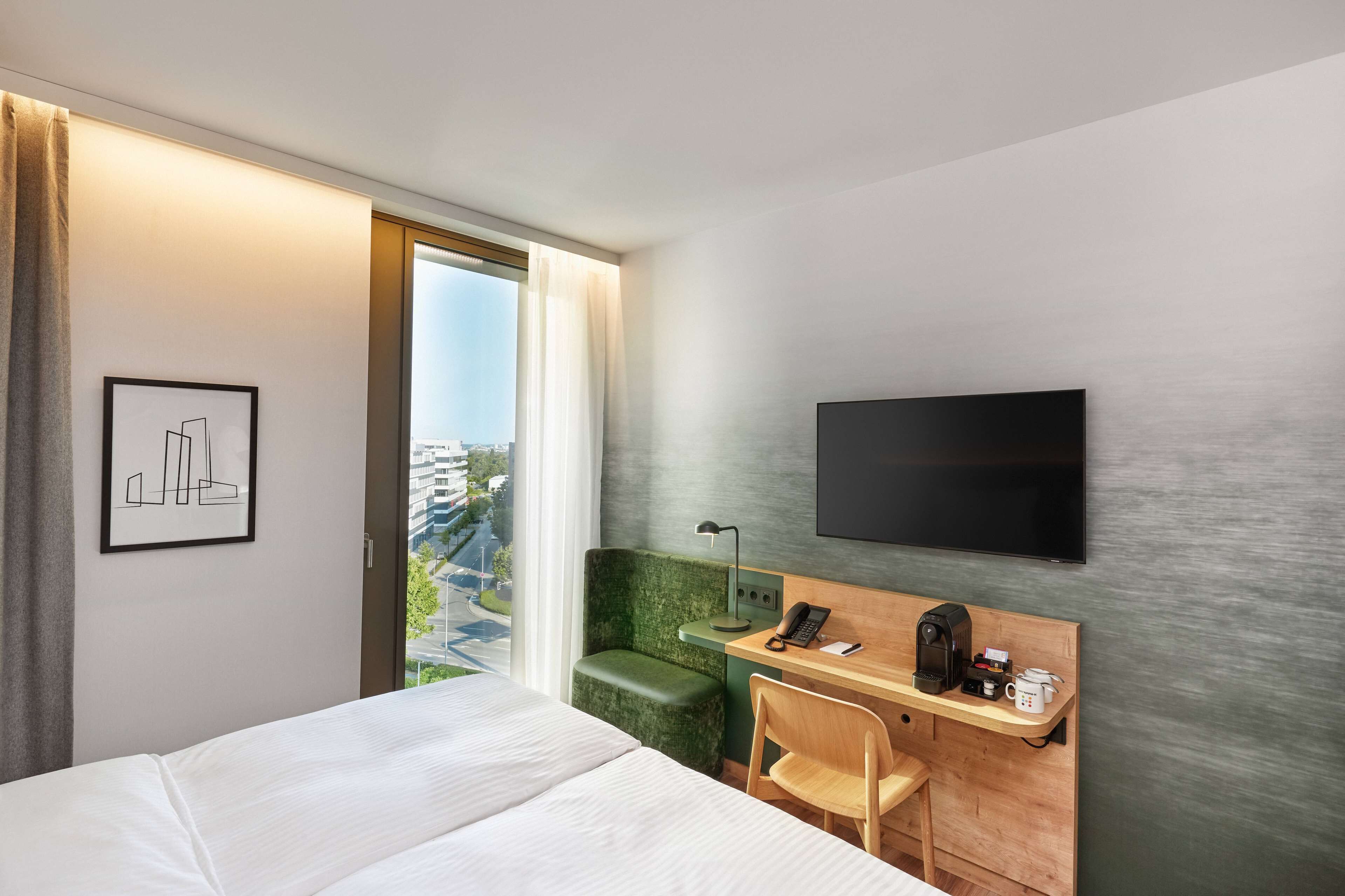 Habitación de hotel con cama, escritorio con silla, televisión de pantalla plana, cafetera y ventana con vistas a edificios.