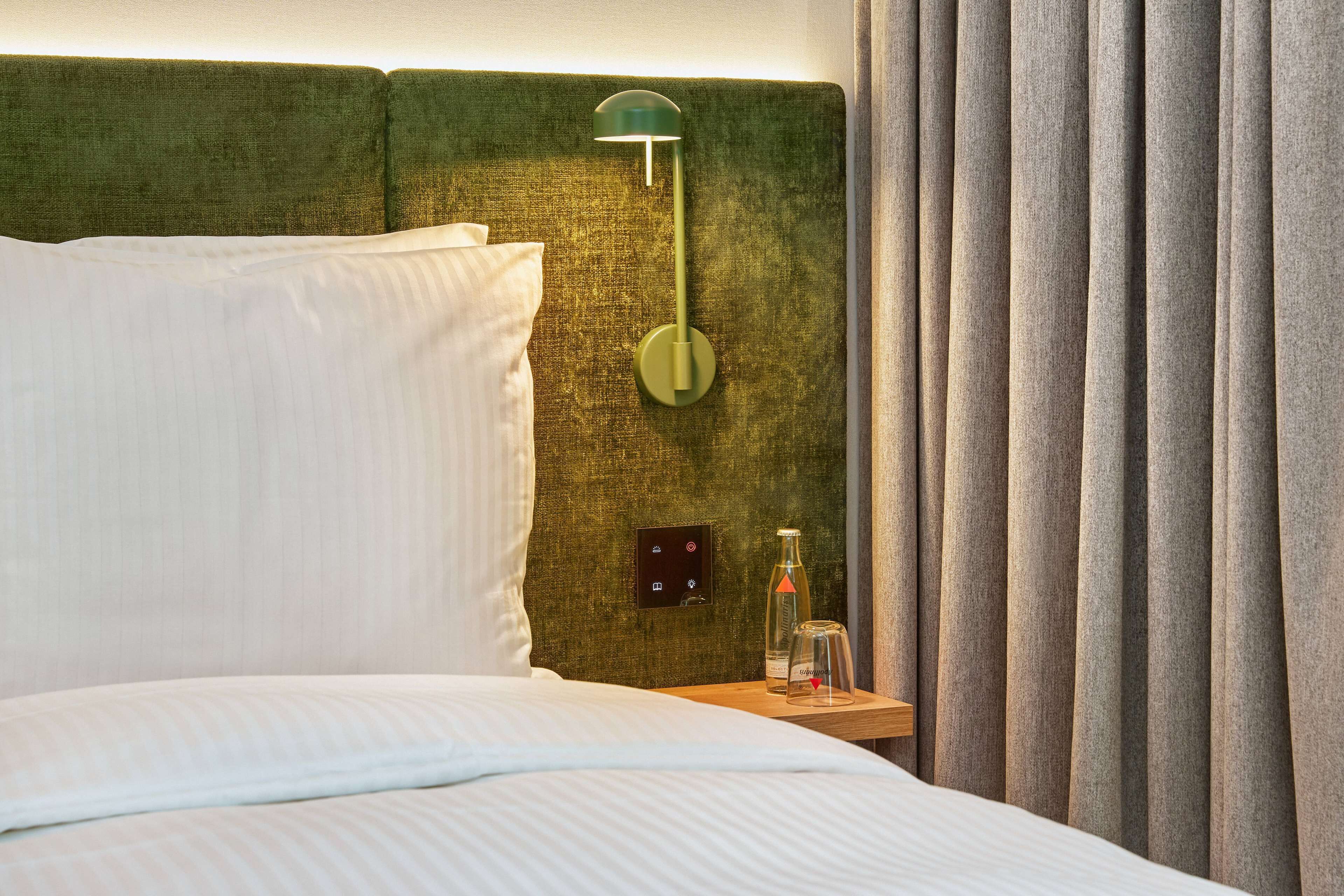 A neatly made bed with white linens, a green headboard, a bedside table with a bottle and glass, and a wall-mounted light fixture.