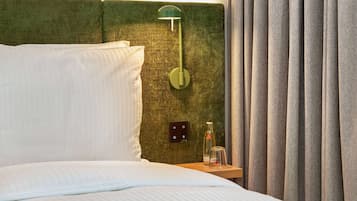 Una cama bien tendida con sábanas blancas, cabecera verde, mesita de noche con una botella y vaso, y una lámpara fijada a la pared.