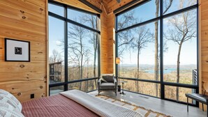 A bedroom with a large bed, a chair, and a view of a forest through large windows. - Blue Ridge Ultra Luxury with Stunning Mountain & Lake Views, Near Aska & Marina (Blue Ridge)