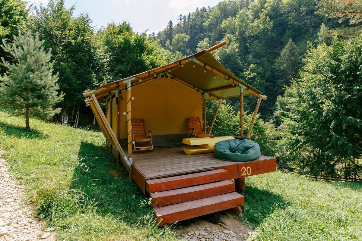 Una pequeña cabaña de una sola habitación con una terraza de madera, dos sillas, un banco y un techo similar a una tienda de campaña.