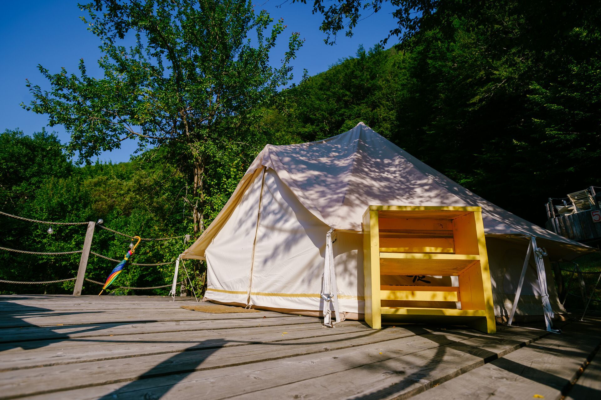 A large tent with an open entrance, surrounded by a wooden deck and ropes, set in a forested area.