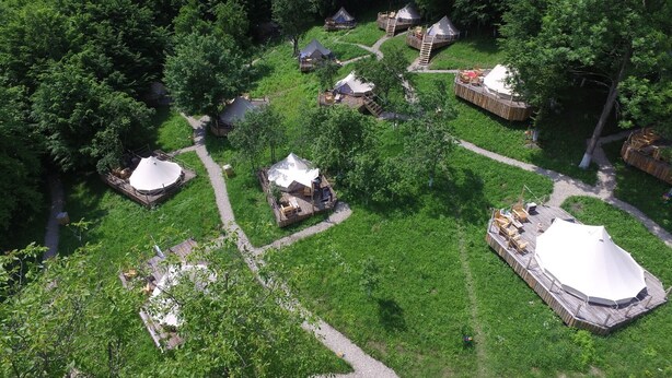 Ein Luftbild zeigt einen Campingplatz mit mehreren Zelten und Holzbauten, umgeben von dichtem Grün.
