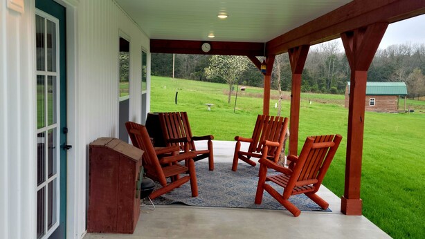 A covered porch with wooden chairs and a small wooden table, a clock, and a view of a green landscape. - Lake Cabin near the Bourbeuse River (Leslie)