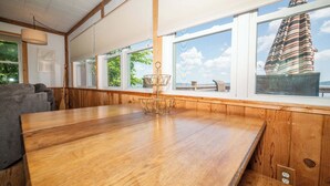 A wooden dining table with a metal basket centerpiece, large windows with a view of the sea, and a gray sofa in a room with wooden paneling. - Waters Edge Cottage (Merrimac)