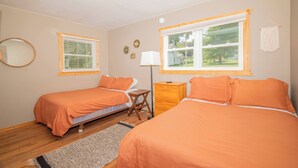 A bedroom with two beds, a nightstand, a lamp, and a window with blinds. - Waters Edge Cottage (Merrimac)