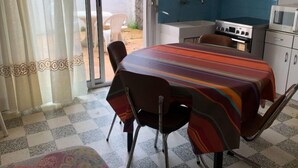 A dining area with a table covered by a striped tablecloth, chairs, a kitchen with blue tiles, and a sliding glass door leading to an outdoor area. - Pretty F2 1 to 4 persons 2 (L'Aiguillon-la-Presqu'île)