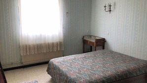 A bedroom with a bed, a small table, a radiator, and a window with curtains. - Pretty F2 1 to 4 persons 2 (L'Aiguillon-la-Presqu'île)