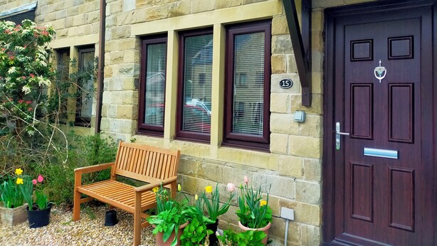 Un bâtiment en pierre avec un banc en bois, des plantes en pot et une porte de couleur brun foncé ornée d’une plaque portant un numéro.